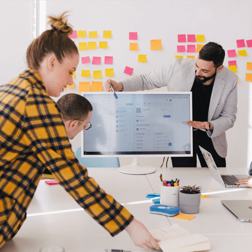 Three coworkers collaborate around a computer monitor displaying a digital dashboard, with colorful sticky notes on the wall behind them.