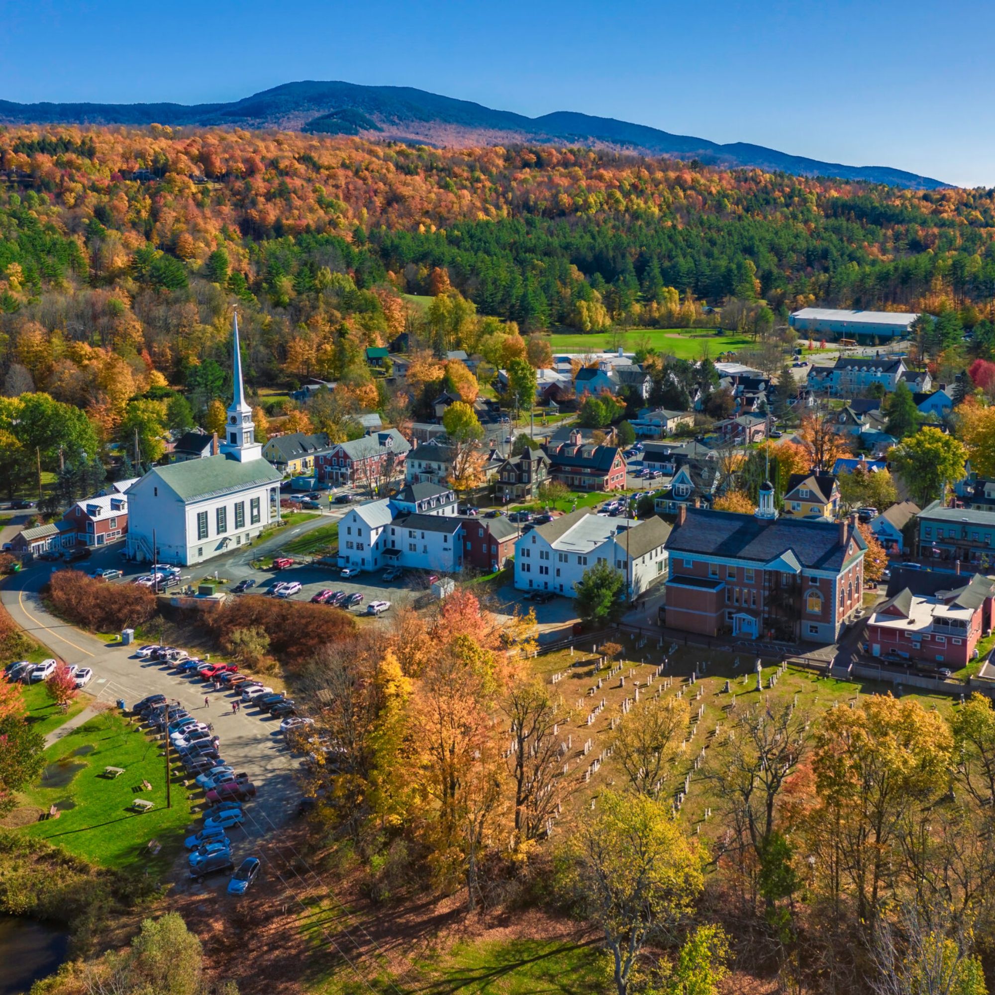 Aerial view of a small New England town in autumn, with a white church steeple, historic brick and clapboard buildings, a cemetery, and surrounding hills covered in fall foliage.
