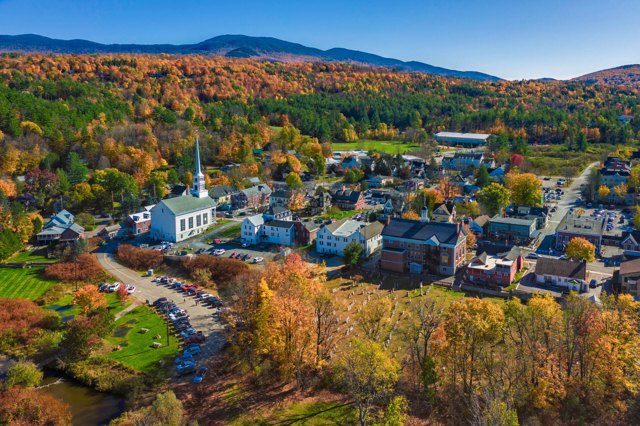 Aerial view of a small New England town in autumn, with a white church steeple, historic brick and clapboard buildings, a cemetery, and surrounding hills covered in fall foliage.