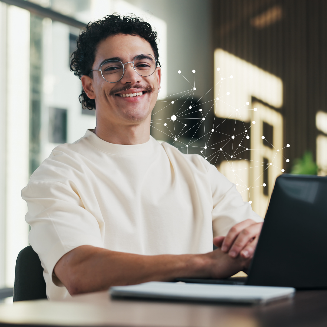 A smiling man wearing glasses works on a laptop at a desk, with a digital network graphic overlaid in the background.