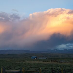 Rural Wyoming landscape with ranch buildings beneath dramatic sunset clouds and distant mountains.
