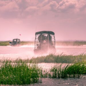 Two airboats traveling through the Florida Everglades, surrounded by marsh grass and open water under a cloudy sky.