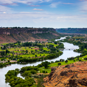 Scenic view of the Snake River winding through a lush green valley in Idaho, surrounded by rugged canyon cliffs, rolling hills, and a partly cloudy blue sky.