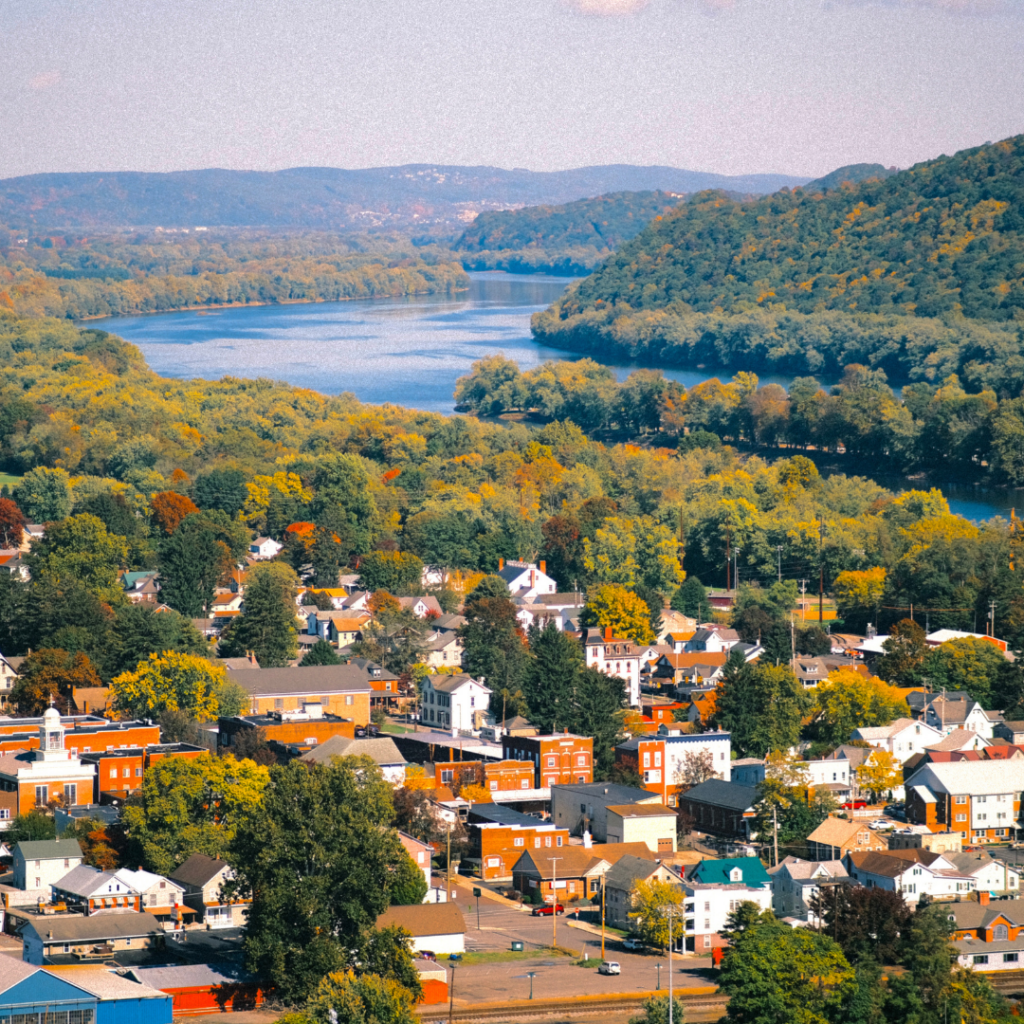Aerial view of a small town surrounded by colorful fall foliage, with a winding river, forested hills, and mountains stretching into the distance.