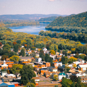 give me alt text Aerial view of a small town surrounded by colorful fall foliage, with a winding river, forested hills, and mountains stretching into the distance.