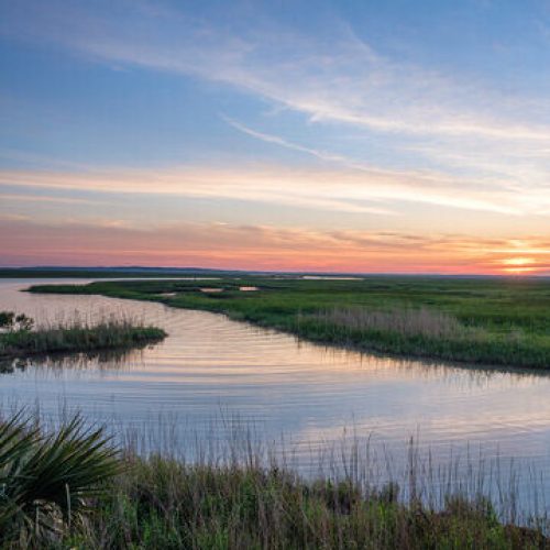 Calm tidal marsh with winding creek under a colorful sunset sky.