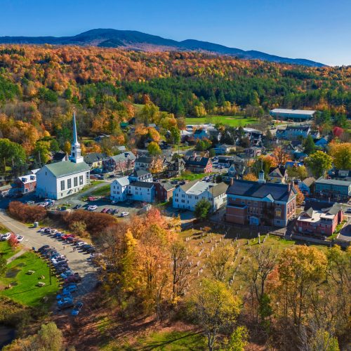 Aerial view of a small New England town in autumn, with a white church steeple, historic brick and clapboard buildings, a cemetery, and surrounding hills covered in fall foliage.